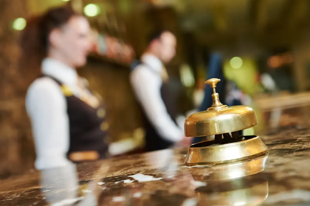 A brass service bell on a hotel reception desk, in sharp focus, with hotel staff members slightly blurred in the background.